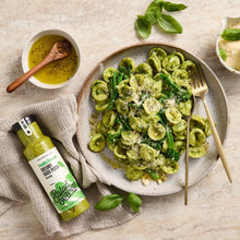 Load image into Gallery viewer, A styled food scene showing a plate of green pasta with broccoli and parmesan, alongside a bottle of Salad Obsessed creamy pasta sauce, a bowl of olive oil, and fresh basil on a light stone surface.