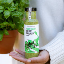 Load image into Gallery viewer, A hand holding a bottle of Salad Servers Freshly Dressed Creamy Basil Pesto dressing, with a potted basil plant in the background.