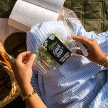 Load image into Gallery viewer, Person holding a salad container with a fork, sitting outdoors with an open book.