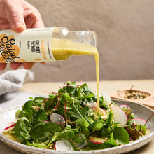 Load image into Gallery viewer, A hand holds a bottle of Salad servers Creamy French Dressing, pouring the golden yellow dressing over a fresh green salad with radishes on a ceramic plate.