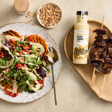 Load image into Gallery viewer, A styled food scene on a light beige surface: a large plate of salad with grilled sweet potato, greens, dressing, and pomegranate, a bottle labeled Salad Servers creamy dressing, and grilled skewers on a wooden board.