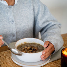 Load image into Gallery viewer, Person holding a bowl of creamy mushroom soup with sliced mushrooms, with a candle beside it.