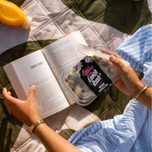 Load image into Gallery viewer, Person holding a container of salad with a fork, next to an open book on a quilted surface.