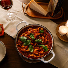 Load image into Gallery viewer, A white enameled cast-iron pot filled with pasta and red sauce, garnished with fresh green basil leaves, set on a wooden table with a wine glass, cheese, and grater in the background.