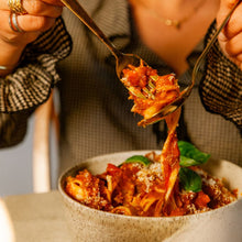 Load image into Gallery viewer, Delicious bowl of pasta in white ceramic bowl. The pasta and ragu sauce is getting put onto a fork by a woman in a black checked shirt.