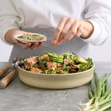 Load image into Gallery viewer, Japanese Salmon & Avocado salad in a bowl on a benchtop with a woman's hands sprinkling on sesame seeds