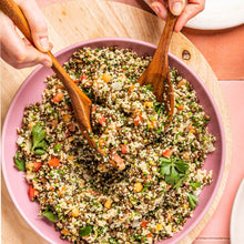Load image into Gallery viewer, A pink serving bowl filled Lentil Tabbouleh and hands holding wooden salad servers picking up a scoop from it.