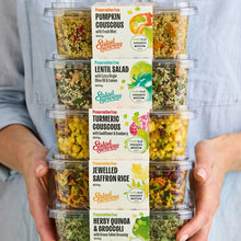 Load image into Gallery viewer, A person holding a stack of five 600g salads. in ascending order Pumpkin Cous Cous with Fresh Mint, Lentil Salad with Extra Virgin Olive Oil, Turmeric Cous Cous with Cauliflower and Cranberry, Jewelled Saffron Rice and Herby Quinoa and Broccoli with Green Tahini Dressing.