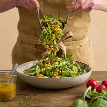 Load image into Gallery viewer, A rustic serving bowl filled with Turmeric Cous Cous and rocket leaves being tossed by a person in a tan coloured apron. The bowl is on a wooden table with a bunch of radishes and a tub of honey mustard dressing on the side.