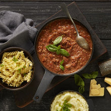 Load image into Gallery viewer, A cast iron pan of Bolognese sauce and a bowl of Fusilli pasta sit on a dark wooden chopping board. A small bowl of mashed potatoes garnished with parsley and a chunk of parmesan sit off to the side.