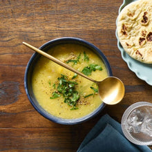 Load image into Gallery viewer, A bowl of chicken and corn soup sits on a wooden table with a plate of flat bread off to the side.