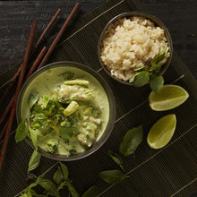 Load image into Gallery viewer, A bowl of Thai Green Chicken Curry and a bowl of rice sit on a dark bamboo mat scattered with wooden chop sticks, lime wedges and mint leaves.