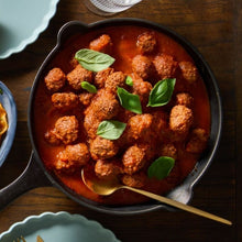 Load image into Gallery viewer, A cast iron pan full of meatballs and garnished with basil leaves sits on a wooden table