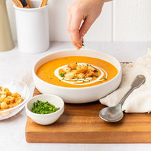 Load image into Gallery viewer, A bowl of pumpkin soup sits on a wooden chopping board in the process of being garnished with croutons and chives. A linen napkin with a silver spoon and bowls of chives and croutons sit off to the side.
