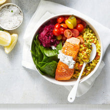 Load image into Gallery viewer, A salad bowl featuring Turmeric Cous Cous, halved cherry tomatoes, baby spinach and picked red cabbage topped with a grilled salmon fillet and yoghurt sauce.