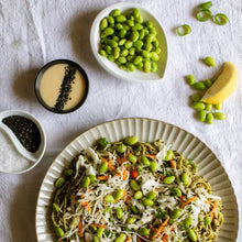 Load image into Gallery viewer, Noodle salad with edamame, sesame seeds, and a small bowl of sauce on a white surface.