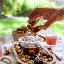 Load image into Gallery viewer, A hand pours granola from a small clear container into a larger clear container labeled 'OATWEIGHTS OAT CHIC & COCOA PUFFER' on a wooden tray, set on an outdoor table with drinks and a salad bowl in a sunny garden setting.