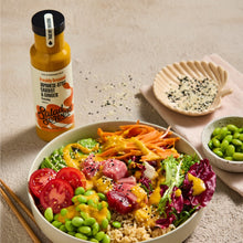 Load image into Gallery viewer, A styled overhead food scene showing a large bowl of salad with rice, edamame, tomatoes, greens, shredded carrots, and pinkish-red slices, alongside a bottle of Salad Senpai dressing and small side dishes on a beige tabletop.