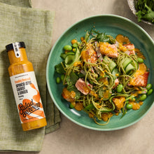 Load image into Gallery viewer, A bottle of Salad Creations freshly dressed Japanese-style carrot & ginger dressing sits next to a green bowl filled with a salad of spaghetti, cucumber, edamame, and salmon on a beige tabletop.