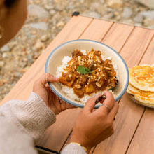 Load image into Gallery viewer, Person holding a bowl of Massaman beef curry with rice, peanuts, and naan bread on a picnic table.