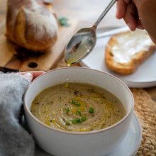 Load image into Gallery viewer, Person drizzling olive oil over a bowl of creamy soup with bread in the background.