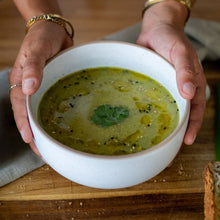 Load image into Gallery viewer, Person holding a bowl of green vegetable soup topped with herbs and olive oil.