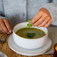 Load image into Gallery viewer, Person garnishing a bowl of green soup with fresh herbs beside a candle and bread.