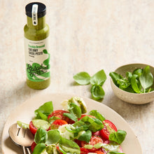 Load image into Gallery viewer, A bottle of Salad servers Freshly Drizzled Creamy Basil Pesto stands next to a plate of fresh basil salad with tomatoes and avocado, with a small bowl of basil leaves on a light stone surface.
