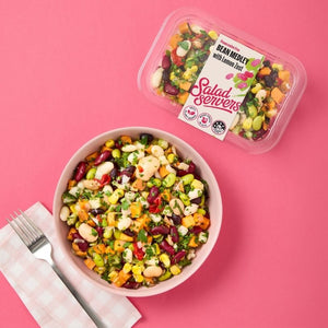 Colourful bean medley salad in a bowl with a container labeled 'Salad Servers' on a pink background.