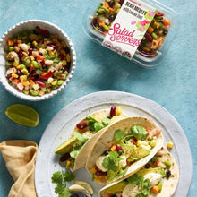 Load image into Gallery viewer, Tacos with salad on a plate, next to a bowl of bean medley salad and a container labeled &#39;Salad Servers&#39;.
