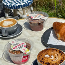 Load image into Gallery viewer, A styled breakfast table scene outdoors featuring a cup of latte with heart latte art, a croissant on a plate, and two clear plastic containers of Sweetbites high protein breakfast boxes on a light stone table.
