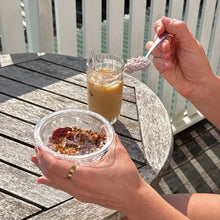 Load image into Gallery viewer, A person holding a clear plastic bowl of granola and cereal on a wooden table, with a glass of iced coffee and a spoon in the background on an outdoor deck.

