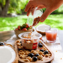 Load image into Gallery viewer, A hand pours granola into a clear plastic cup of overnight oats on a wooden tray, set on a table outdoors with a blurred green lawn background. Other breakfast items like a strawberry bowl, a glass of pink drink, and a coconut are visible.
