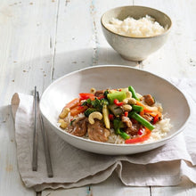 Load image into Gallery viewer, Bowl of stir-fry with vegetables and meat on a white wooden table with a side of rice.
