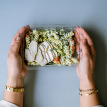Load image into Gallery viewer, Hands holding a clear container of creamy pesto pasta with sliced chicken.
