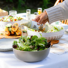 Load image into Gallery viewer, A hand in a cream ribbed sweater pours dressing from a bottle labeled 'Bourgeon Salad Dressing' into a bowl of mixed green salad on an outdoor dining table.
