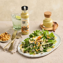 Load image into Gallery viewer, A bottle of Salad Seasoning creamy French dressing stands next to a glass of water with a lime wedge, with a plated salad featuring asparagus, greens, blue cheese, and dressing on a textured beige table.
