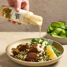 Load image into Gallery viewer, A hand holds a bottle of Secret Sauce Creamy Hummus dressing, pouring a thick beige sauce over a plated bowl of creamy hummus with broccoli, olives, and a lemon wedge, with a side of sliced cucumbers in the background.
