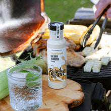 Load image into Gallery viewer, A bottle of Salad Servers creamy hummus dressing stands on a wooden serving board outdoors near a grill, with a glass of iced drink and grilled food in the background.

