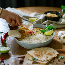 Load image into Gallery viewer, Person dipping a piece of flatbread into a bowl of rice and curry on a wooden table.
