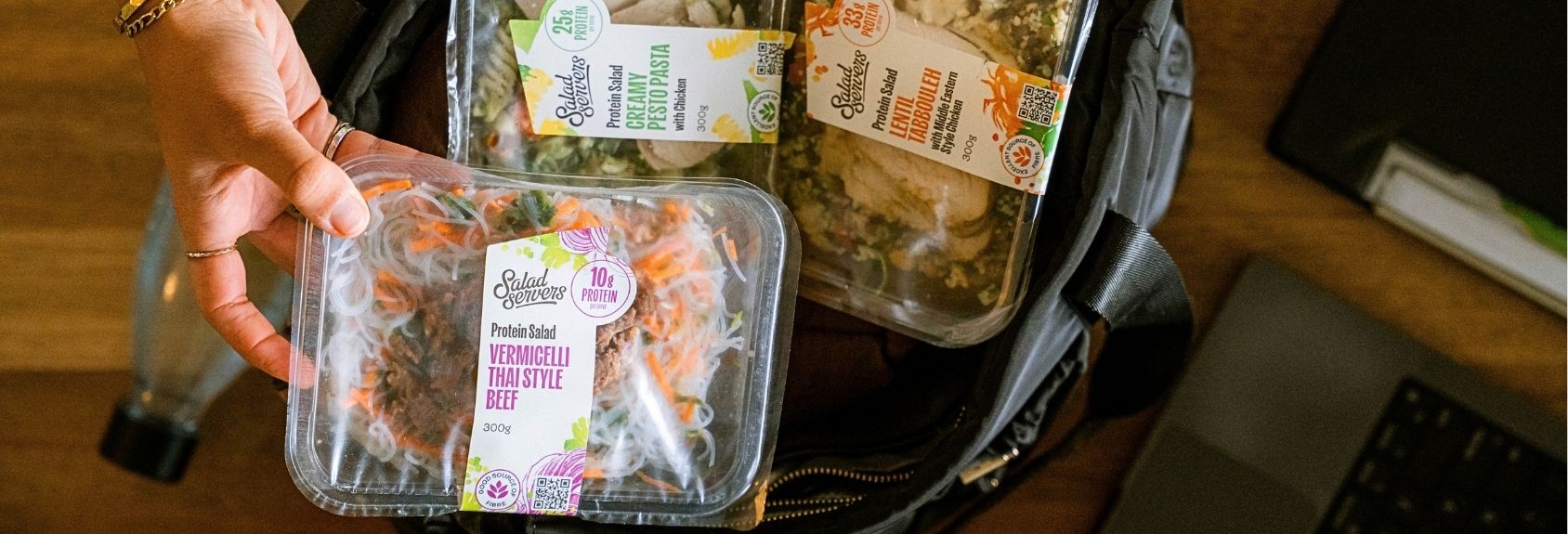 Hand packing a Salad Servers Vermicelli Thai Style Beef protein salad into a bag with Creamy Pesto Pasta with Chicken and Lentil Tabbouleh with Middle Eastern Style Chicken, ready to grab and go.