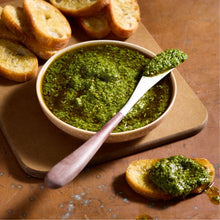 Load image into Gallery viewer, Bowl of green pesto with a spoon, surrounded by crostini on a wooden board.
