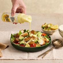 Load image into Gallery viewer, A hand holds a bottle of dressing above a green salad bowl on a table runner. The salad contains chicken, greens, tomatoes, and avocado, with a creamy yellow dressing being poured over it.
