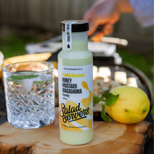 Load image into Gallery viewer, A bottle of Salad Servers Honey Mustard Macadamia dressing stands on a wooden slice outdoors, with a glass and a lemon beside it. A blurred person is visible in the background near a grill.
