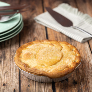 Apple pie in a metal pie dish on a wooden table with plates and a knife in the background.