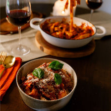 Load image into Gallery viewer, A bowl of pasta with red sauce and fresh basil leaves in the foreground, with another bowl of pasta and a glass of red wine blurred in the background on a dark wooden table.
