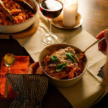 Load image into Gallery viewer, A bowl of pasta with tomato sauce and fresh basil leaves on a dining table, with a hand holding a fork and another hand resting near the bowl. A glass of red wine, a candle, and other table setting details are visible in the background.
