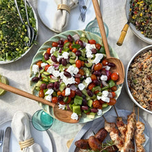 Load image into Gallery viewer, Greek Salad on a busy table of lots of yummy food with wooden salad servers
