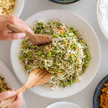 Load image into Gallery viewer, Person using wooden utensils to mix a bowl of noodles with vegetables on a light gray surface.
