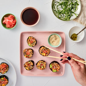 Sushi rolls on a pink plate with a hand holding chopsticks, surrounded by soy sauce, wasabi, and pickled ginger.